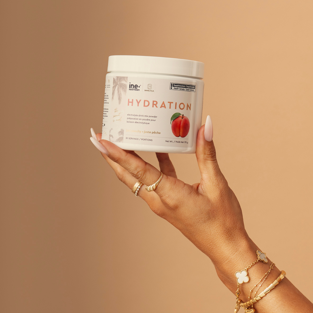 A hand with long manicured nails holds a white tub of Eho Hydration drink mix against a solid warm beige background. The tub features a white lid and a white label with a red strawberry graphic.